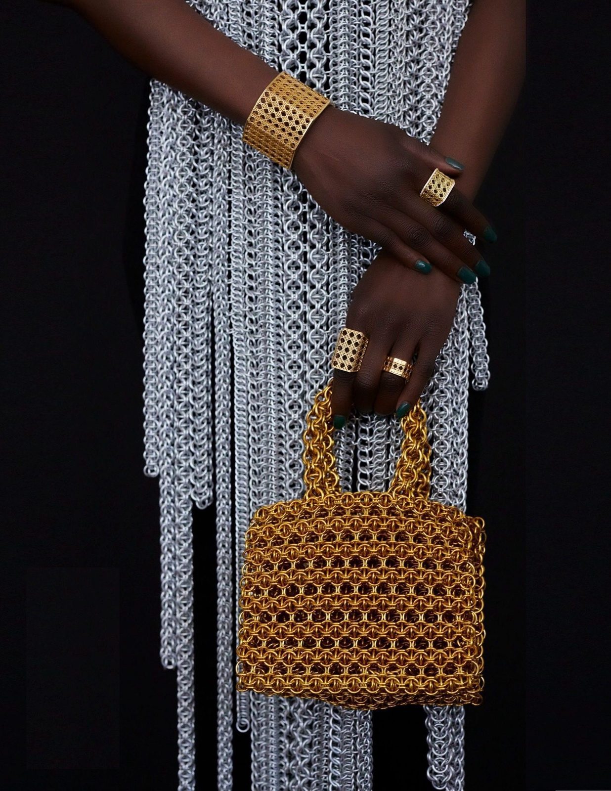 The Accessories Council first jewelry edition cover model photographed in dark studio light wearing the Free Maison Aluminum Apega custom couture piece — a full-length silver hand-woven chainmail fringe dress — while holding the gold anodized aluminum Free Maison Nowy Bag with two top handles. Her hands are adorned with gold chainmail rings and a wide gold chainmail cuff bracelet.