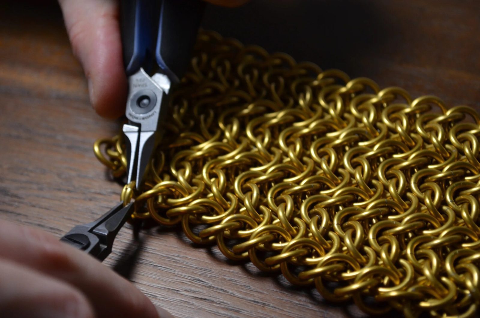 Close-up of hands using steel-nosed pliers to open and weave individual gold anodized aluminum rings into a classical chainmail panel on a wooden workbench. The dense European 4-in-1 weave pattern fills the frame, showing the hand-crafting process behind every Free Maison piece.