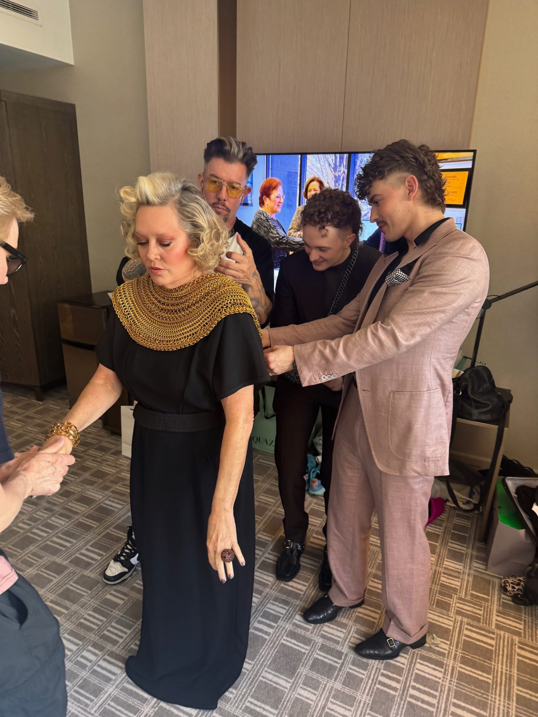 Jesse Aviv and Tay Dun fitting a custom gold anodized aluminum Free Maison chainmail Mane piece on costume designer Molly Rogers backstage before The Devil Wears Prada 2 New York City premiere. Rogers stands in a black dress as the two founders carefully adjust the piece around her.