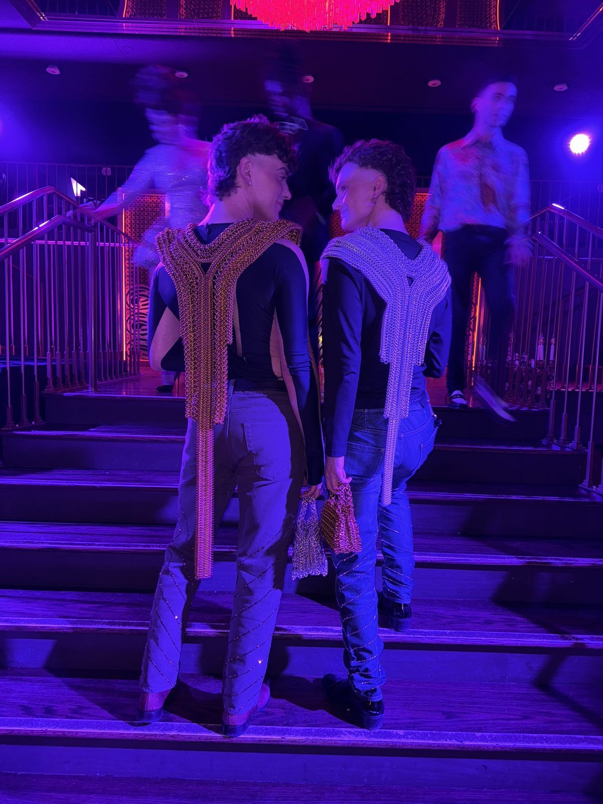 Free Maison co-founders Jesse Aviv and Tay Dun at the Susanne Bartsch NYE party in Times Square, New York City, bathed in purple and red stage lighting. Jesse wears the Free Maison gold Merovingian lame chainmail piece and Tay wears the silver Persephone lame chainmail piece. Both carry Free Maison Nowy Mini Totes in gold and silver anodized aluminum.