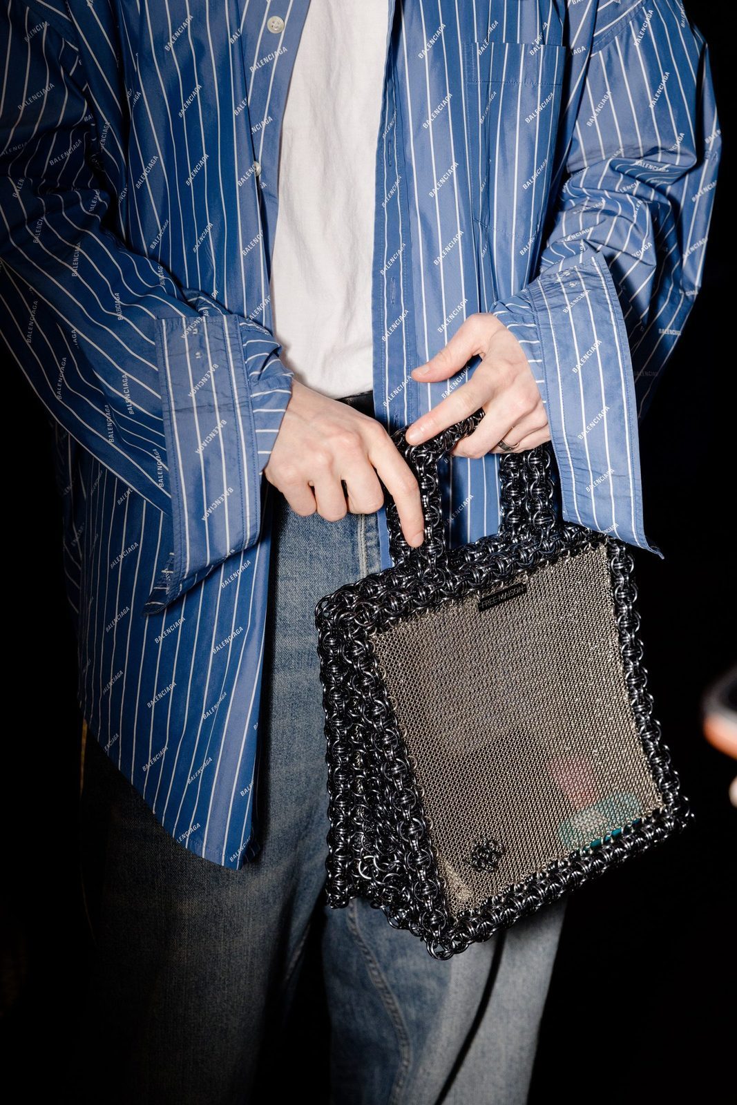 A person wearing a blue pinstripe blazer holds the Free Maison Cyborg Sentinel — a structured hand-woven chainmail tote in stainless steel and storm gray anodized aluminum — by its two short top handles at their side against a dark background.