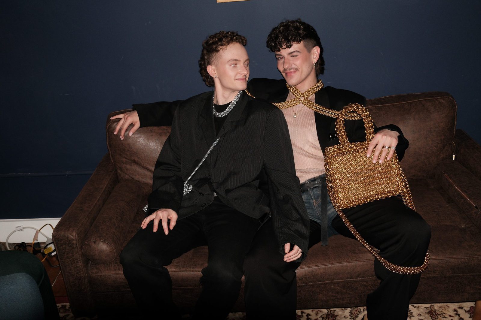 Free Maison co-founders Jesse Aviv and Tay Dun seated together on a brown leather sofa against a navy blue wall. Jesse wears the Free Maison Bit Bag and a chainmail choker. Tay wears the Free Maison Gold Valenciennes Harness and carries the Free Maison Gold Heater Chainmail Bag.