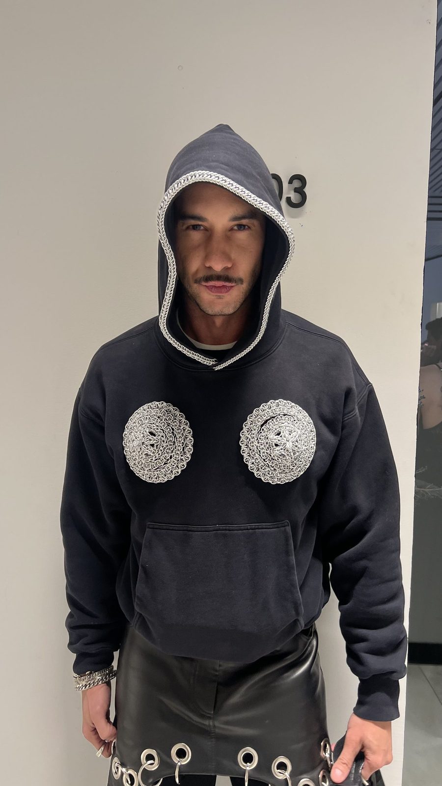Martin Gregory at the Grace Ling opening wearing the Free Maison Artemis Hoodie — a black oversized hoodie featuring a hand-woven silver chainmail cuirass detail on the chest and chainmail trim lining the hood opening, paired with a black leather skirt. Shot against a white exterior wall.