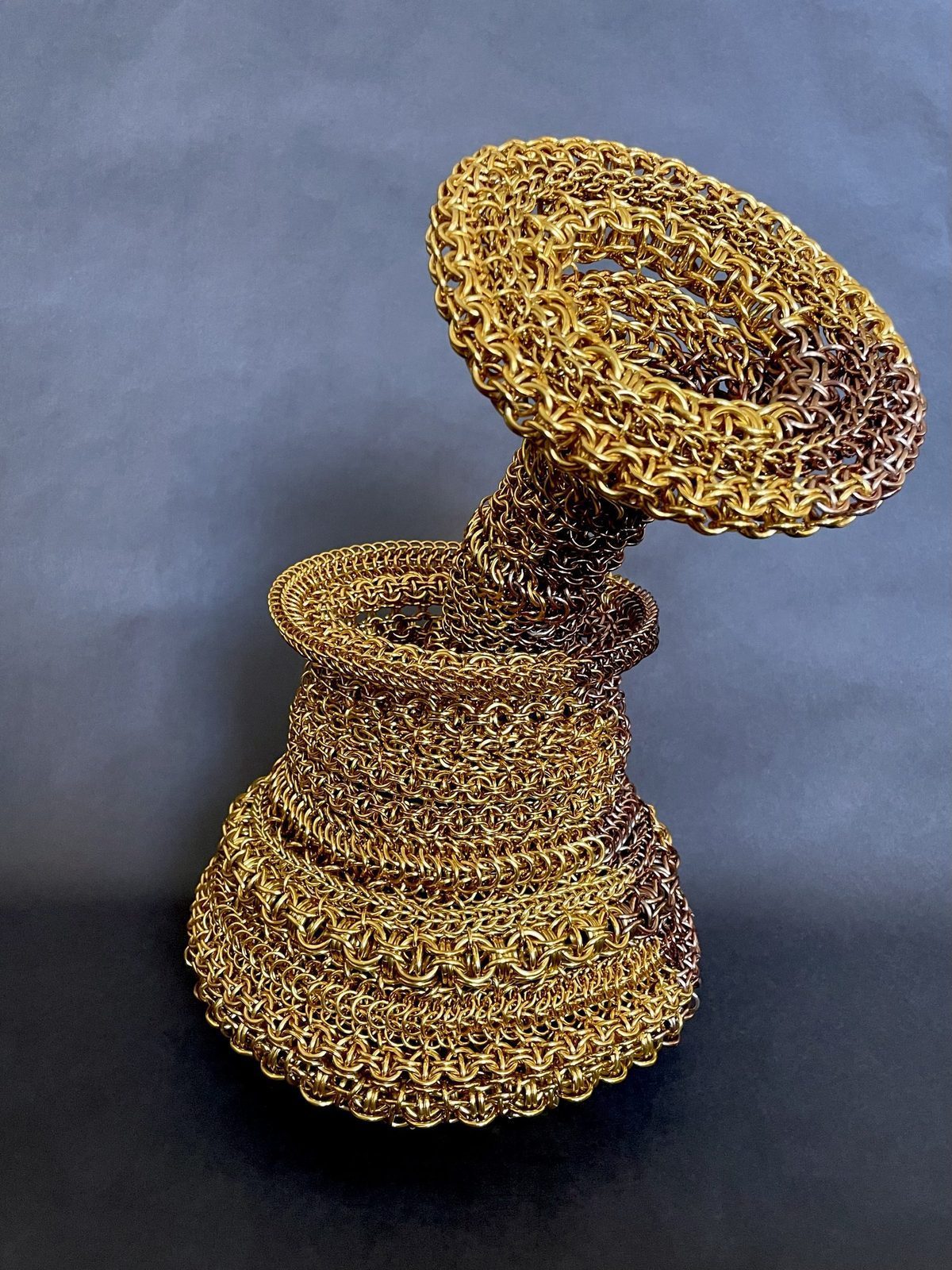 Jesse Aviv's Free Maison chainmail sculpture art piece hand-woven in old penny and gold anodized aluminum. The piece — titled Honey Pot — was featured in Fiber Art Now magazine. The sculptural form spirals upward in stacked rings on a gray background.