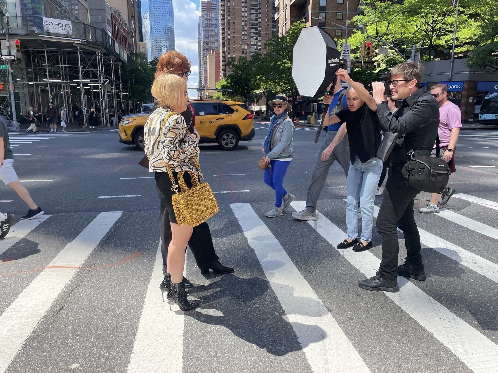 Cindy Gallop on a New York City street photography shoot with photographer Chad Johnson. Gallop poses at a crosswalk carrying the Free Maison Gold Heater Chainmail Bag in gold anodized aluminum. A crew member holds a large softbox light beside her as taxis and pedestrians pass in the background.