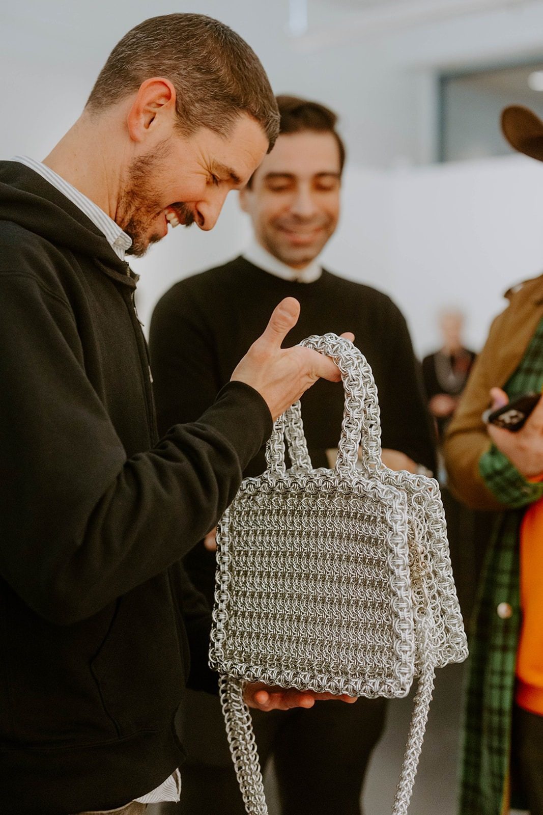 A smiling man holds the Free Maison Silver Heater Chainmail Bag up at eye level to examine it closely at the Free Maison New York Fashion Week 2024 launch event at a gallery in Tribeca, New York City. Other gallery guests are visible in the softly lit background.