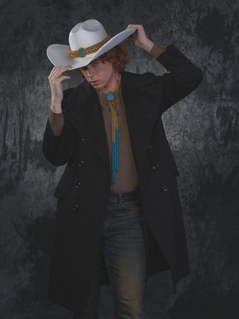 Model Kasey Connor with curly red hair wearing a white wide-brim cowboy hat adorned with a Free Maison gold hand-woven chainmail hatband, and a Free Maison bolo tie in gold chainmail inset with turquoise gemstones. Shot against a neutral background.