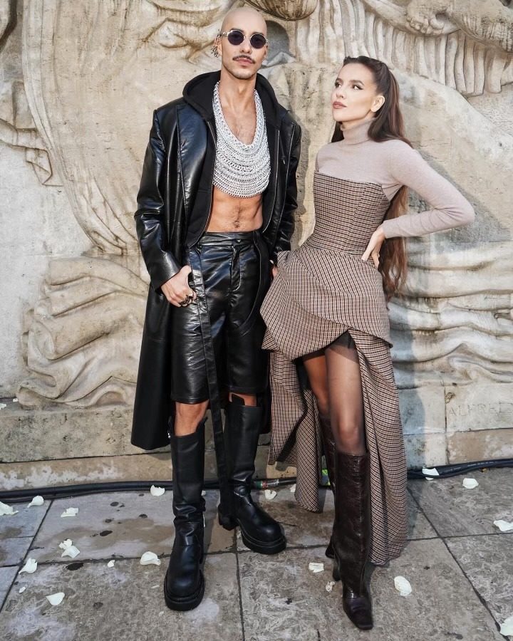 Martin Gregory at the Rick Owens Paris Fashion Week show 2025, standing against a carved stone wall wearing a long black leather coat and a custom Free Maison silver Hanover Mane worn as a bandeau across his chest. Paris Fashion Week attendees pass in the background.