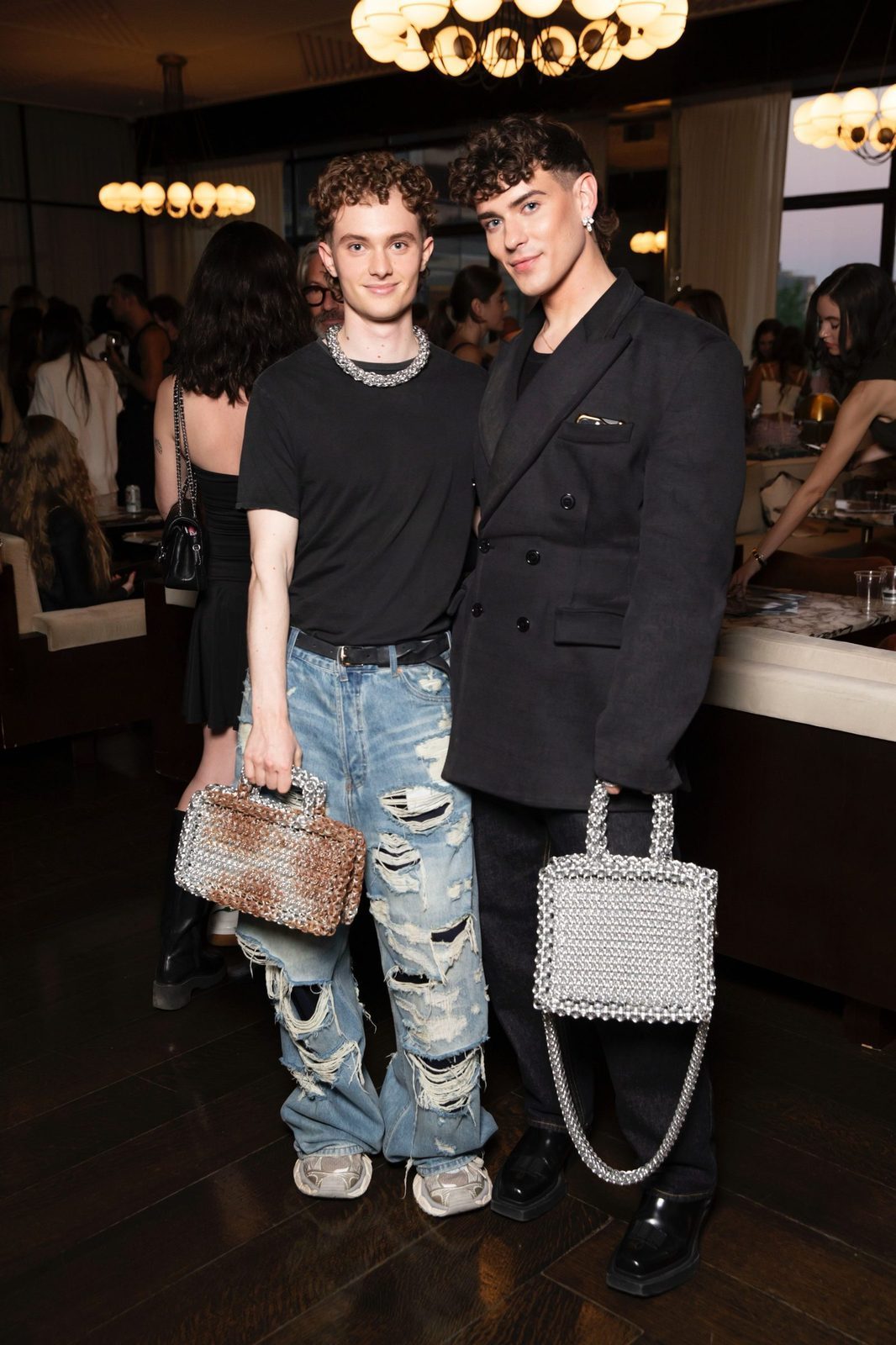 Free Maison co-founders Jesse Aviv and Tay Dun at Milk Studios in New York City. Jesse carries the Free Maison Silver Chainmail Heater Tote and Tay carries the Free Maison chainmail rust bag. Both bags catch the warm ambient light of the room.