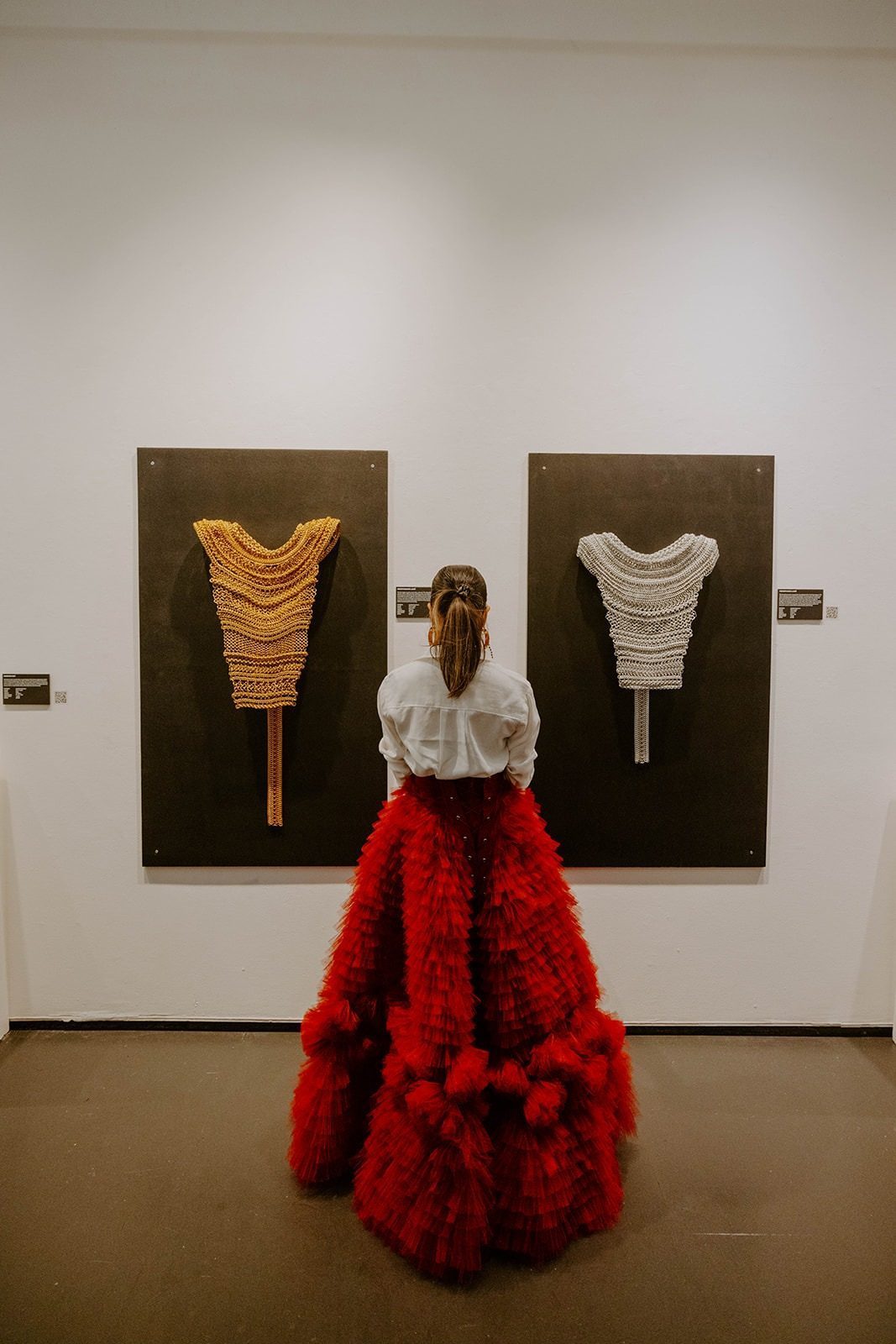 A person in a red ruffled skirt stands viewing the Free Maison award-winning Merovingian Lame and Persephone Lame chainmail garments mounted on a white gallery wall like art at New York Fashion Week. The two hand-woven anodized aluminum pieces are displayed at eye level in the gallery space.