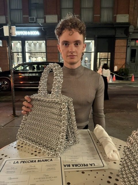 Free Maison co-founder Jesse Aviv seated at an outdoor table at La Pecora Bianca in New York City, holding up the Free Maison Silver Heater Tote in hand-woven anodized aluminum chainmail with both hands, smiling. Other diners and the restaurant terrace are visible behind him.