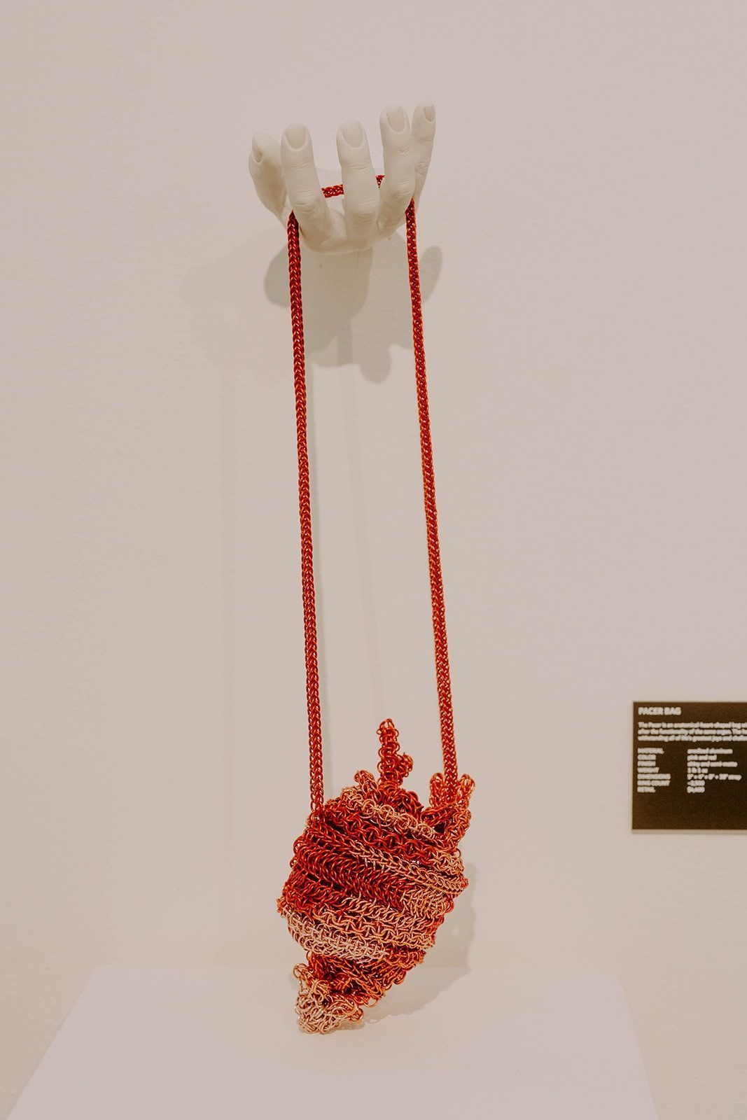 The Free Maison Pacer Heart Bag — an anatomically-shaped hand-woven chainmail bag in red and rose gold anodized aluminum — displayed on a pedestal and mounted on the wall like art at the Free Maison New York Fashion Week 2024 launch event. A small label card is propped beside the mount.