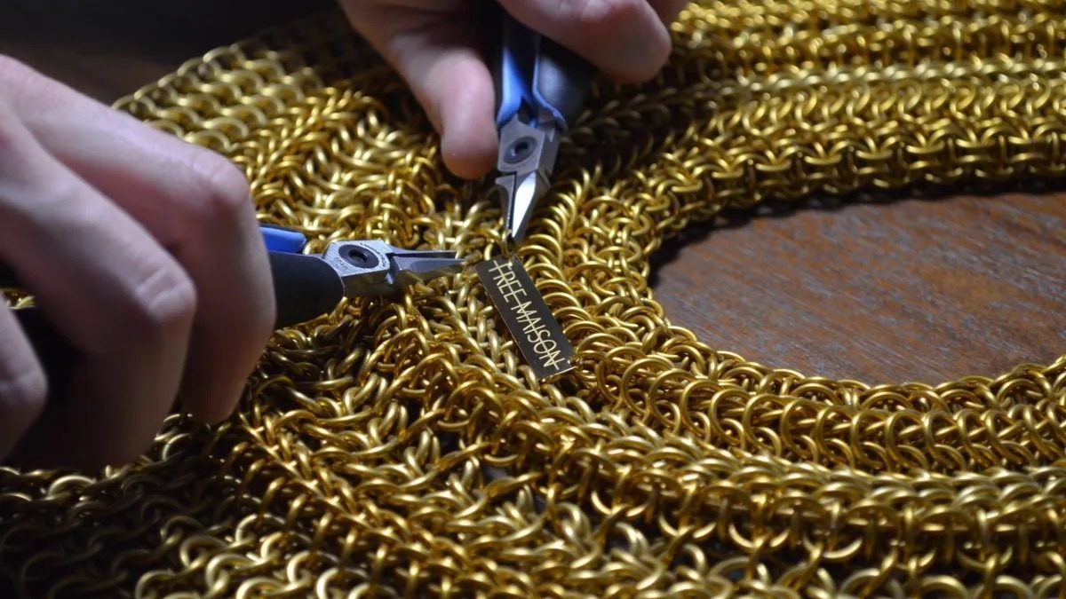 Close-up of Free Maison co-founder Jesse Aviv's hands handcrafting a Free Maison chainmail piece using the house's proprietary ciphered technique, working two pairs of pliers to interlock individual gold anodized aluminum rings. The Free Maison brand tag is being woven directly into the finished work.