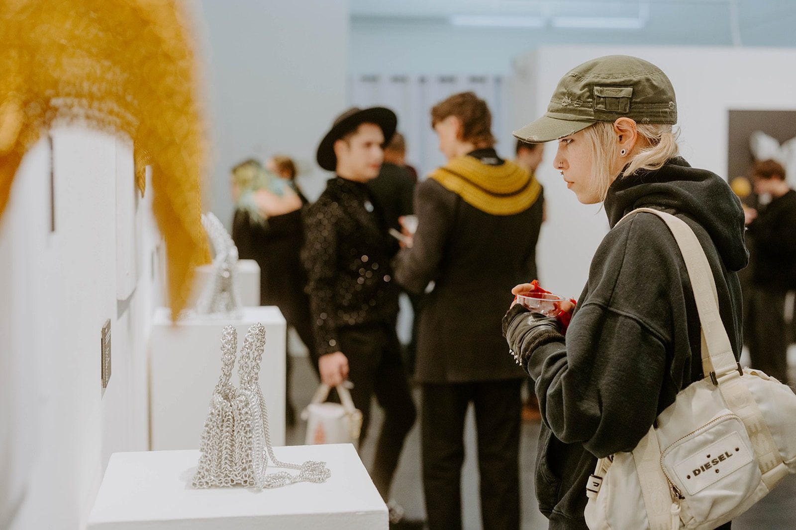 Cheeky MA wearing a green cap leans in closely to examine the Free Maison Silver Naoie Chainmail Bag displayed on a white plinth at the Free Maison New York Fashion Week launch event. The bag catches the overhead gallery lighting as other guests mingle in the background.