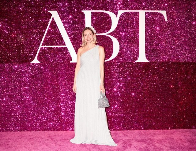 World-renowned astrologer Francesca Vuilleman at the American Ballet Theatre event wearing a white one-shoulder formal gown and carrying the Free Maison Nowy Silver Tote Bag in hand-woven anodized aluminum chainmail. She stands in front of a large pink glitter ABT backdrop as other gala guests in formal attire appear behind her.