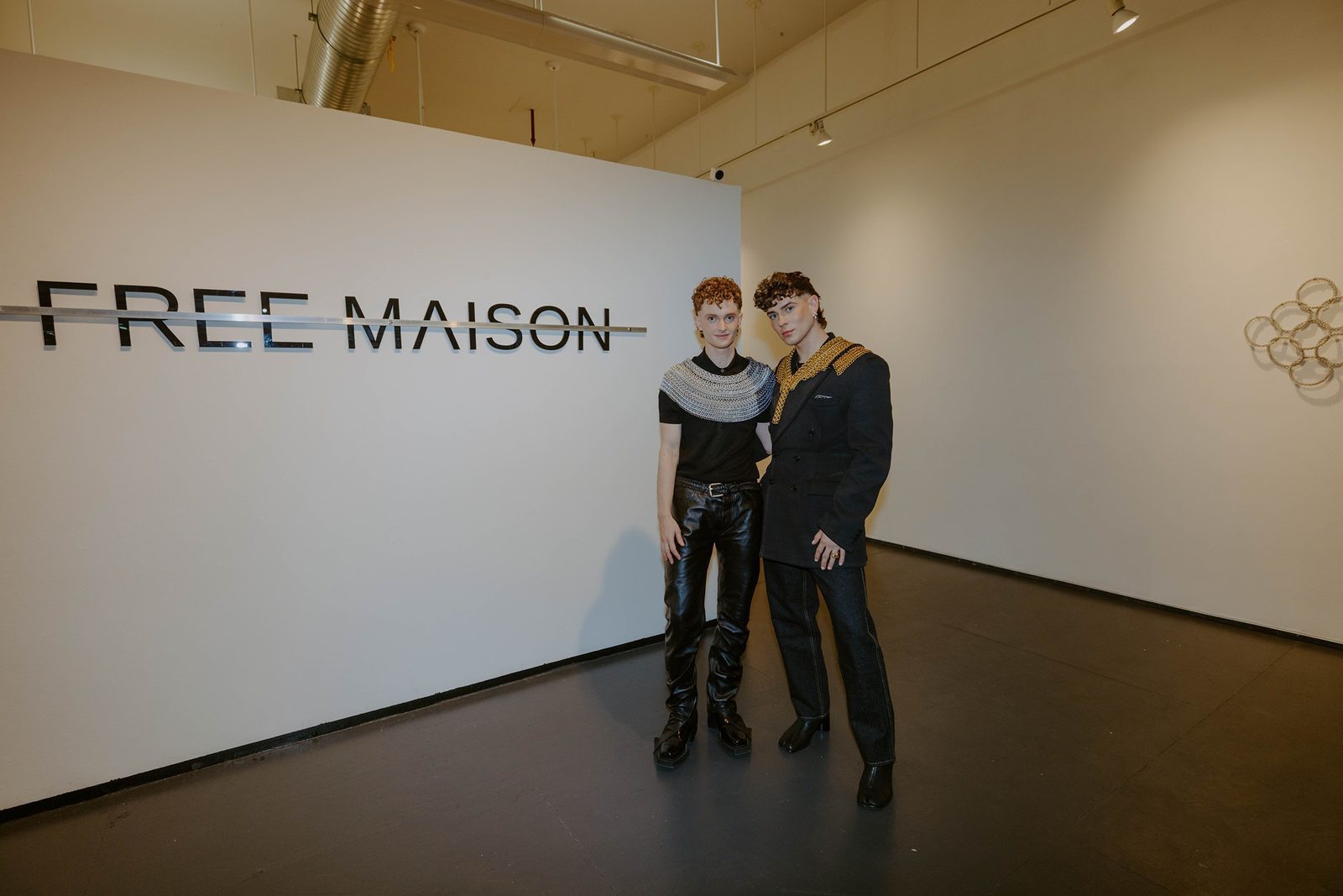 Free Maison co-founders Jesse Aviv and Tay Dun at the Free Maison launch event, standing in front of a large illuminated gallery sign reading 'FREE MAISON.' Jesse wears the Free Maison Hanover Silver Mane and Tay wears the Free Maison Gold Novotny Mane. Hand-woven chainmail art objects are mounted on the gallery walls behind them.